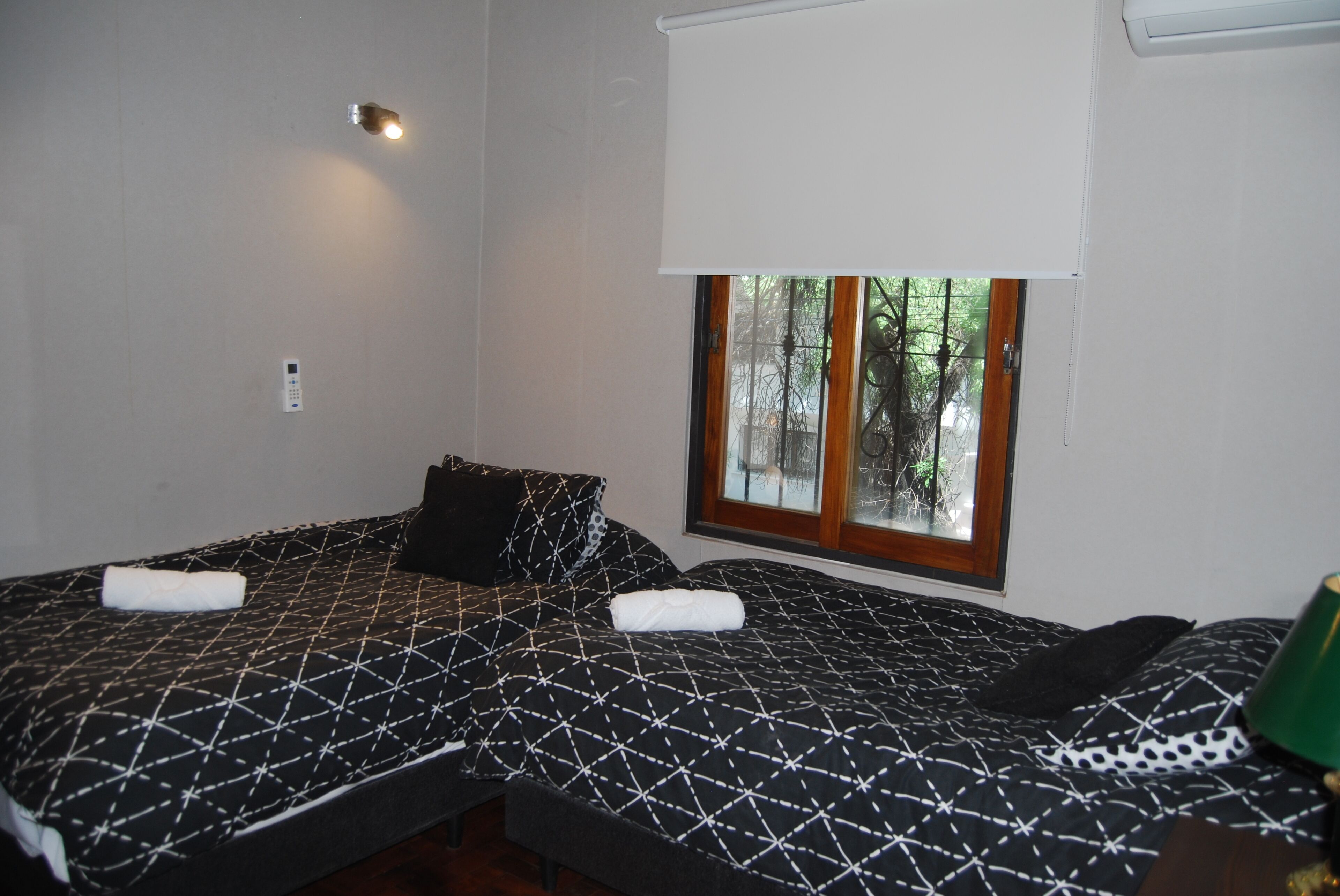 A room with two beds, a window with a view of trees, a wall-mounted light, and a thermostat.