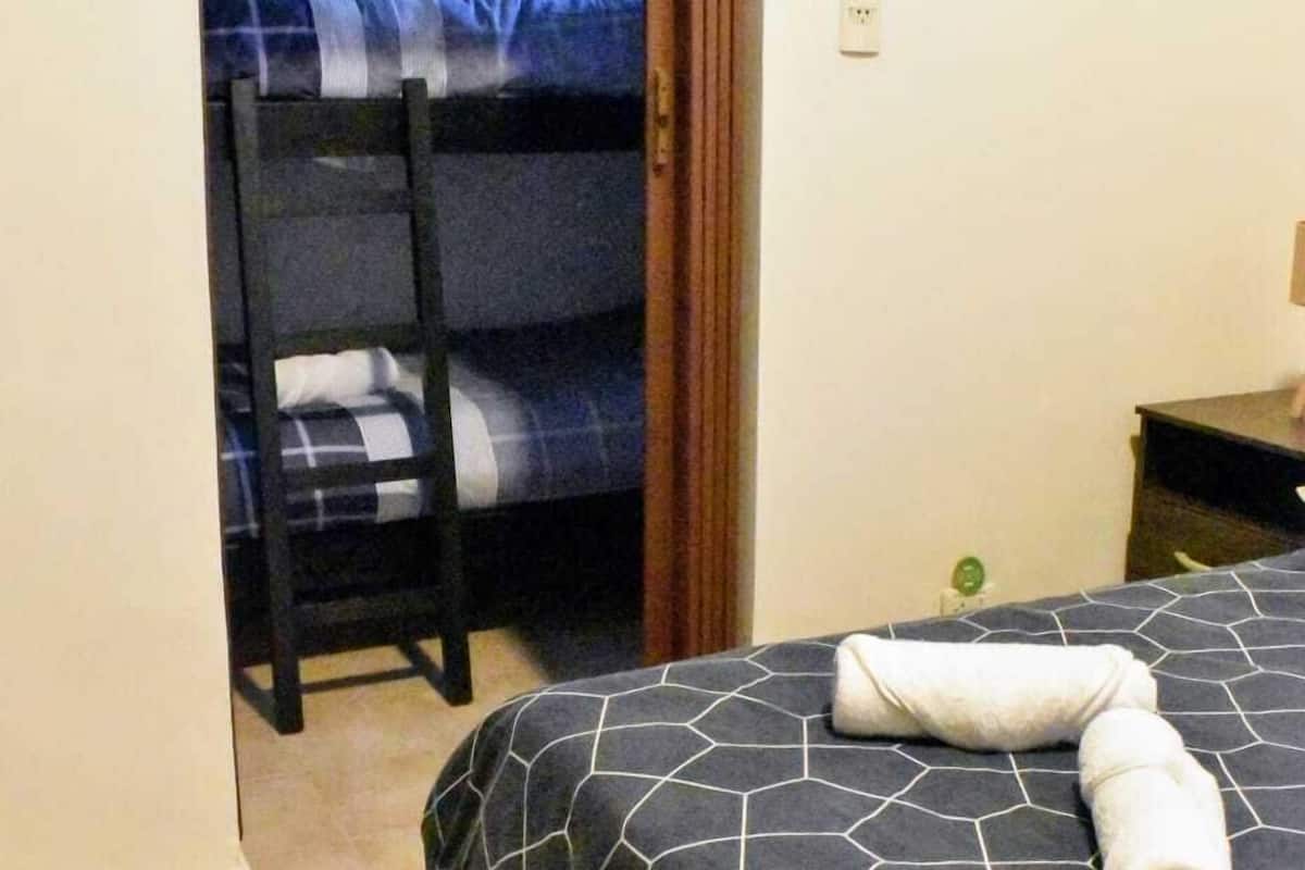 A bedroom with a bunk bed, a bed with a geometric-patterned cover, a rolled towel on the bed, a framed picture on the wall, and a door leading to another room.