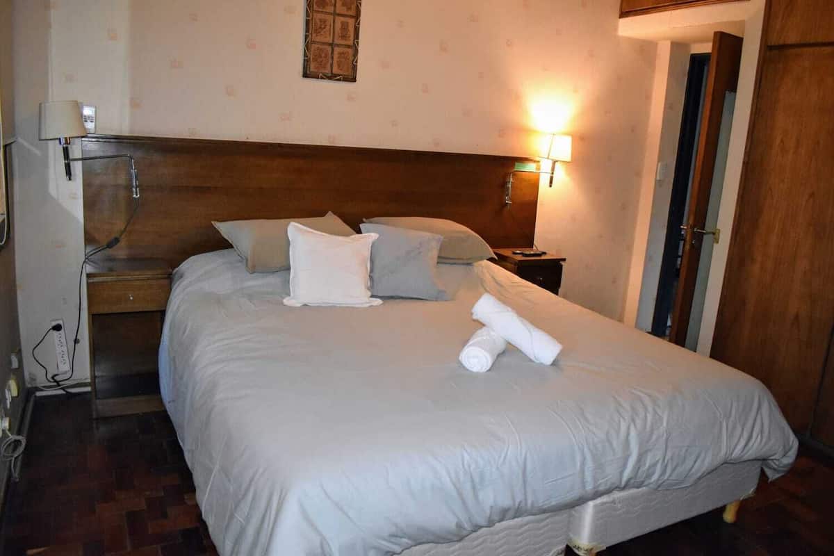 A neatly made bed with a white comforter and pillows, a bedside lamp, and a rolled towel on the bed.