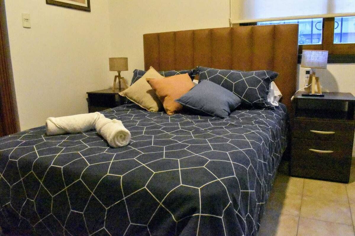 A neatly made bed with a geometric-patterned bedspread, a rolled towel, and several pillows. There is a nightstand with a lamp and a small table with a bottle on it.