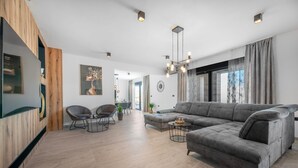 Een ruime woonkamer met een grijze hoekbank, een salontafel en moderne stoelen. Er is een groot raam met gordijnen, een plafondlamp en een aan de muur bevestigde televisie.