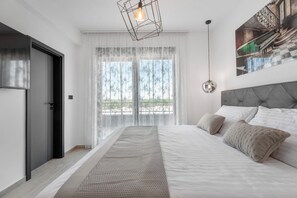 Quarto moderno com uma cama grande, cabeceira cinza e vista para o exterior através de cortinas leves.