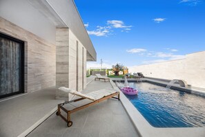 Un área de piscina exterior moderna con una tumbona, una fuente con forma de cisne y un cielo azul despejado.