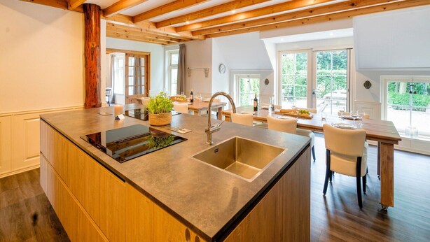 Een moderne keuken met een centraal eiland, houten balken, grote ramen en een eethoek met een tafel en stoelen.