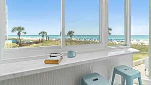 Een kamer met een groot raam dat uitkijkt op een strand, een klein krukje en een boekje op de vensterbank.
