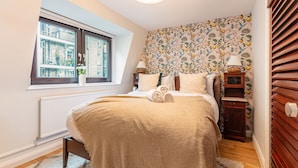 A bedroom with a large window, a radiator, a bed with a beige blanket, a nightstand with a lamp, and a wall with a fruit and bird wallpaper. - Spacious 2BR House by Madame Tussauds  (London)