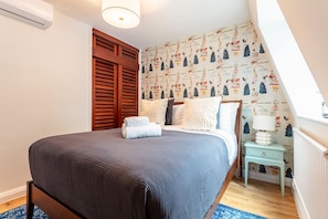 Un dormitorio con una cama grande, contraventanas de madera, un papel tapiz estampado y una mesita de noche con lámpara.