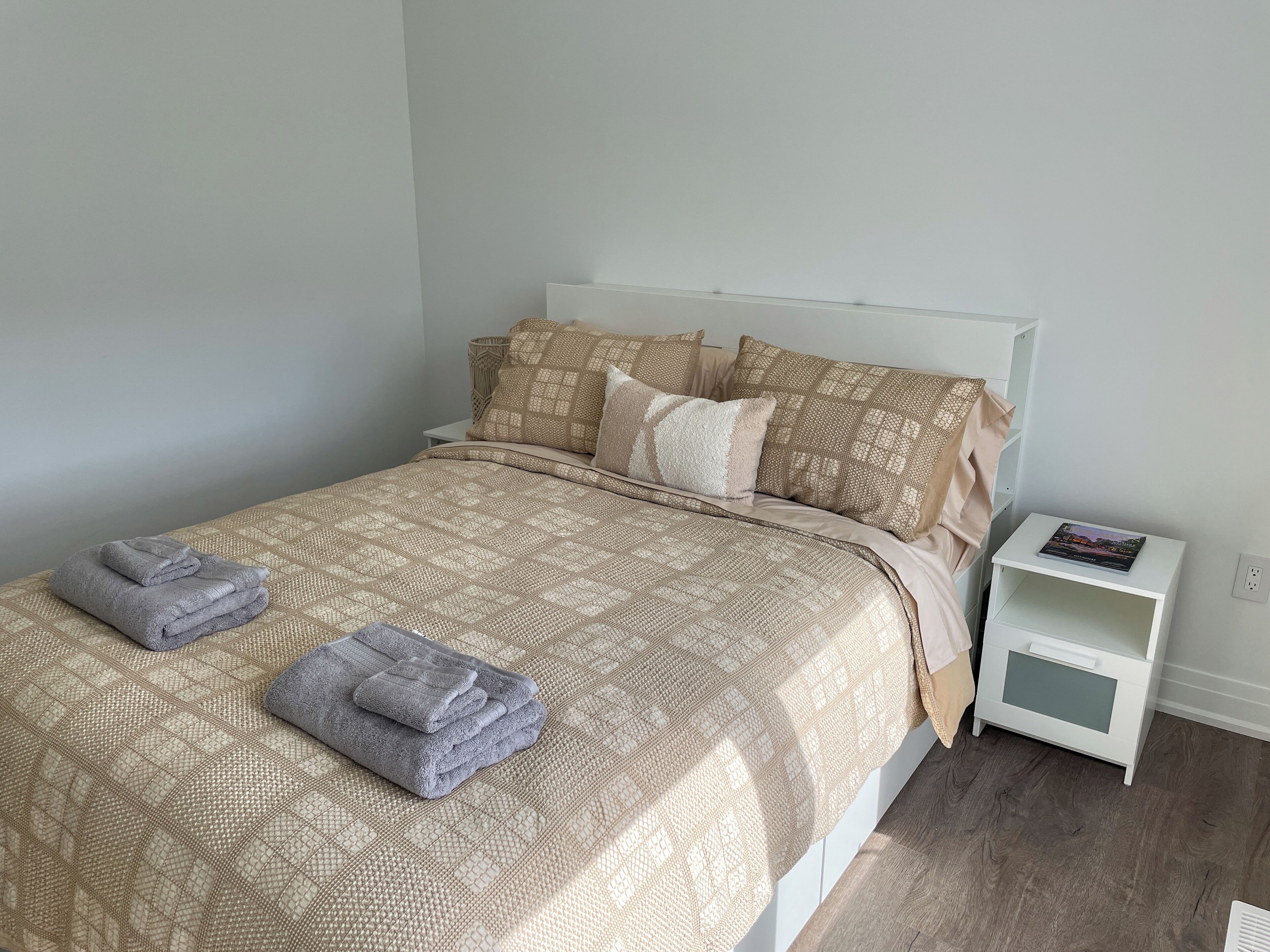 A neatly made bed with a patterned bedspread, two folded towels, and a bedside table with a book.