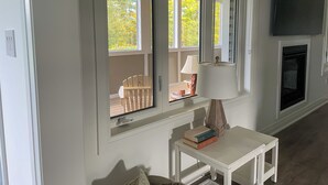 Une pièce dotée d’une grande fenêtre, d’une table blanche avec une lampe et d’un téléviseur à écran plat fixé au mur.
