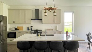 Une cuisine moderne dotée d’un grand îlot central, de tabourets de bar, d’appareils électroménagers en acier inoxydable et offrant une vue sur l’extérieur.