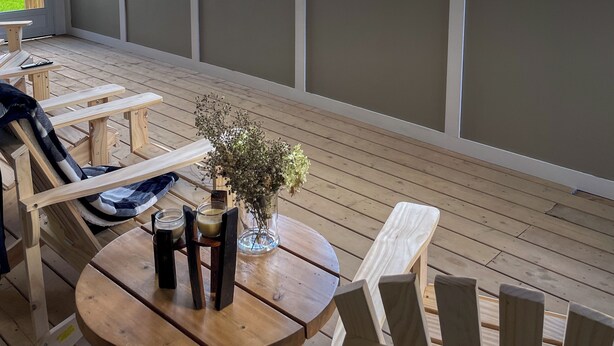 Un espace extérieur aménagé avec des sièges, protégé par un toit, comprenant des chaises en bois et une table ronde, de grandes fenêtres et une vue sur un paysage verdoyant.