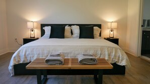 Un lit bien fait, recouvert d’une couvre-lit à rayures, deux tables de chevet avec des lampes et un banc en bois sur lequel sont posées des serviettes.