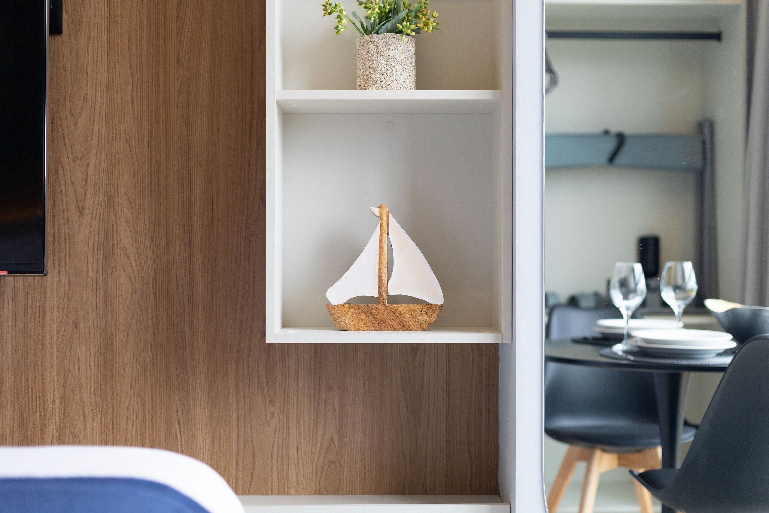 A modern interior with a wooden wall, a white shelf with a small sailboat model, and a dining area with black chairs and a table set for two.
