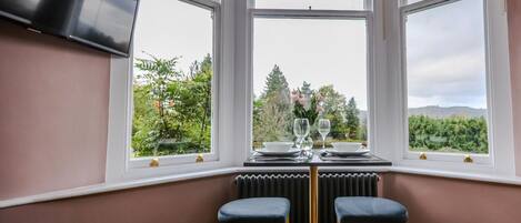 A corner dining area with a table set for two, two chairs, and a view of greenery outside.