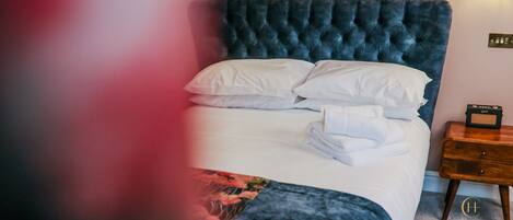 A neatly made bed with white linens and a dark blue headboard. There is a wooden nightstand with a radio beside the bed.