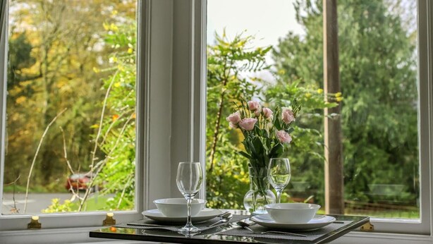 A dining table with a vase of flowers and two wine glasses set for two, positioned by a large window with a view of trees and a road. - Fair Rigg by Cradel Haus (Windermere)