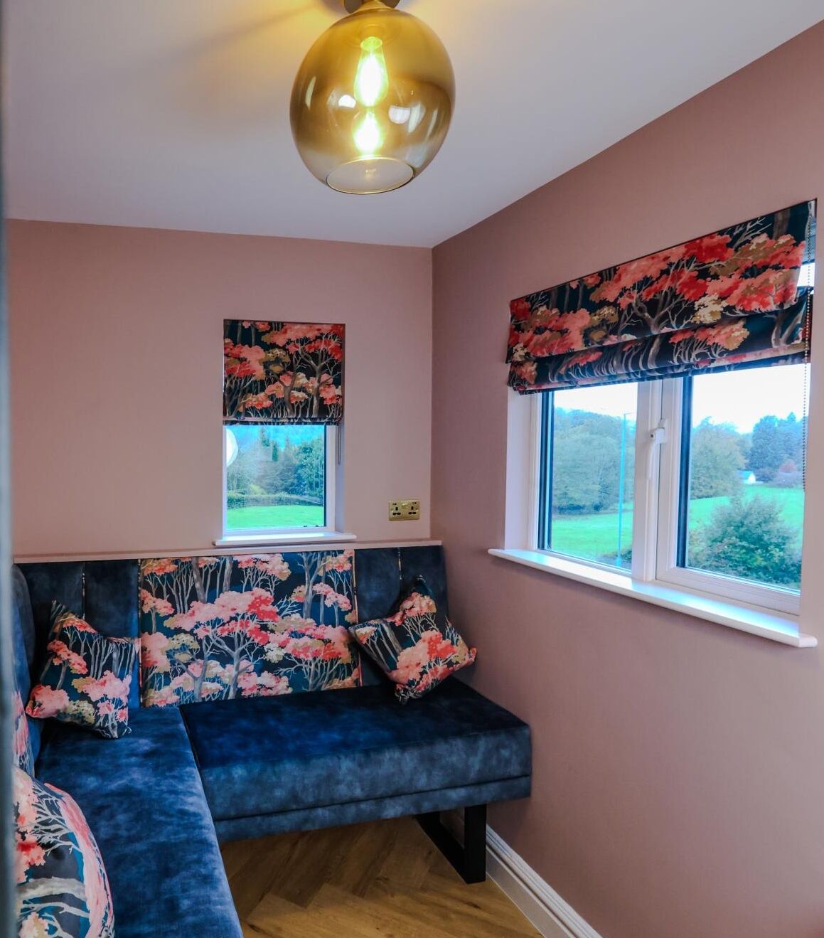 A room with a blue sofa, floral-patterned cushions, a round ceiling light, and a window with a view of greenery.