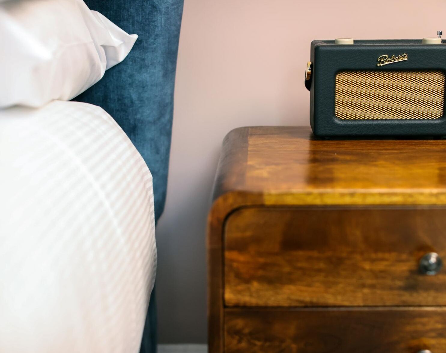 Un lit avec un oreiller blanc et une radio ancienne posée sur une table de chevet en bois.