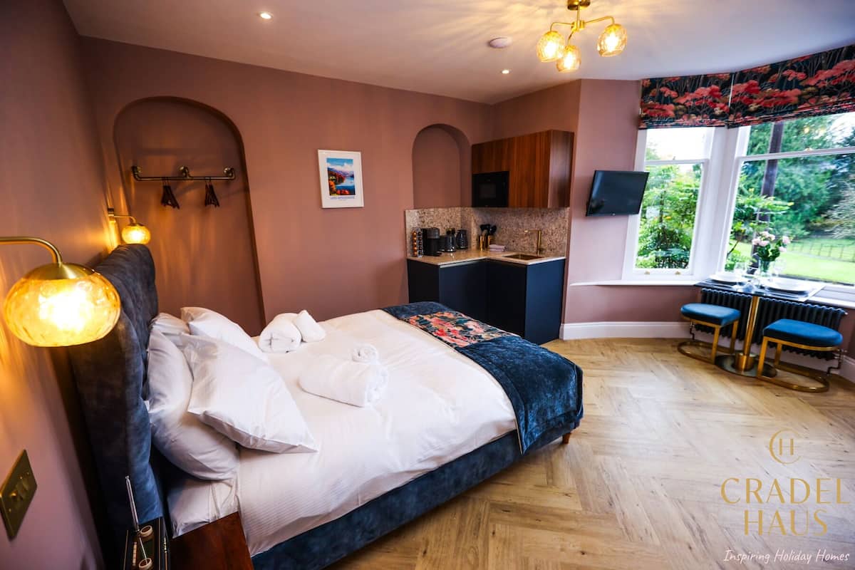 Une chambre à coucher comprenant un lit, des lampes de chevet, une petite kitchenette et une fenêtre donnant sur un espace verdoyant.
