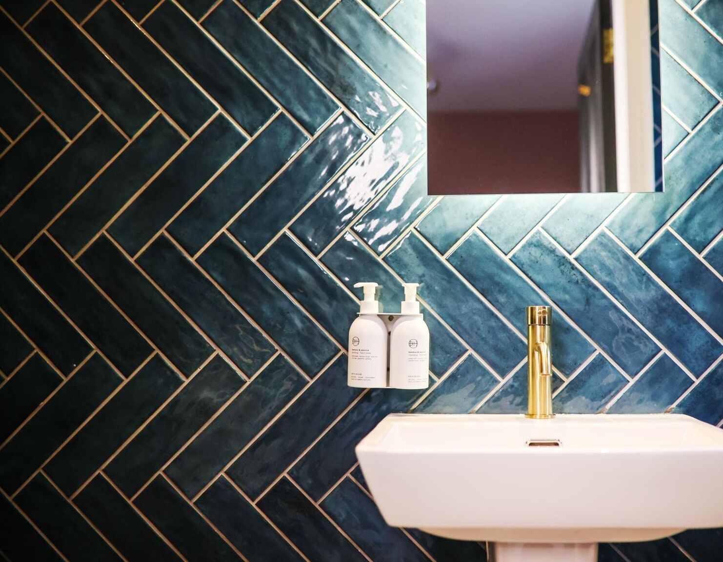 Un baño con salpicadero de azulejos tipo espiga y un lavabo blanco con grifo dorado.