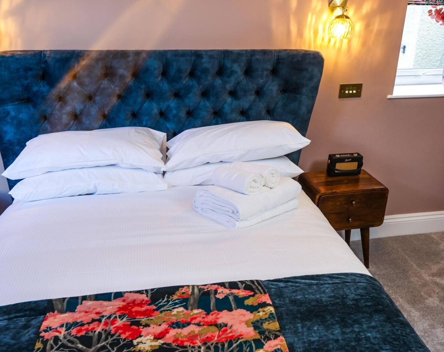 Een net opgemaakt bed met witte lakens en kussens, een blauw gestoffeerd hoofdbord en een nachtkastje met een radio.