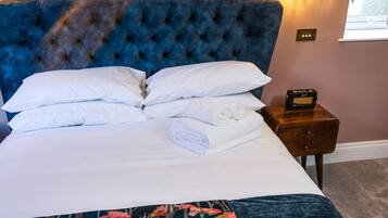 Een net opgemaakt bed met witte lakens en kussens, een blauw gestoffeerd hoofdbord en een nachtkastje met een radio.