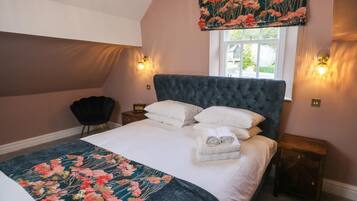 Een slaapkamer met een groot bed, een donkerblauwe hoofdbord, twee nachtkastjes, een stoel en een raam met uitzicht op groen.