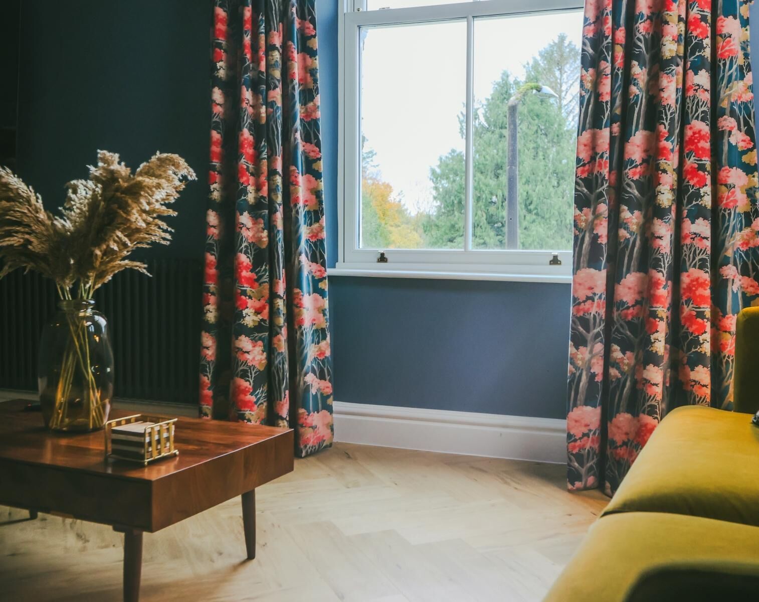 Ein Zimmer mit Fenster, dunklen Wänden, einem Holztisch und blumigen Vorhängen.