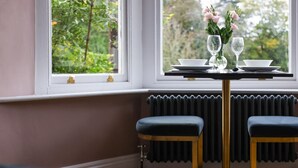 A dining area with a table set for two, two stools, a window with a view of greenery, and a radiator below the window. - Fair Rigg by Cradel Haus (Windermere)
