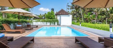 A swimming pool with lounge chairs and umbrellas, surrounded by greenery and a white building.