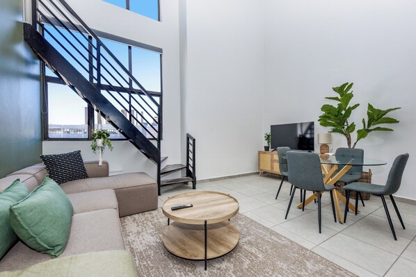 A modern living room with a sofa, a round coffee table, a dining area with chairs, a TV, and a staircase.
