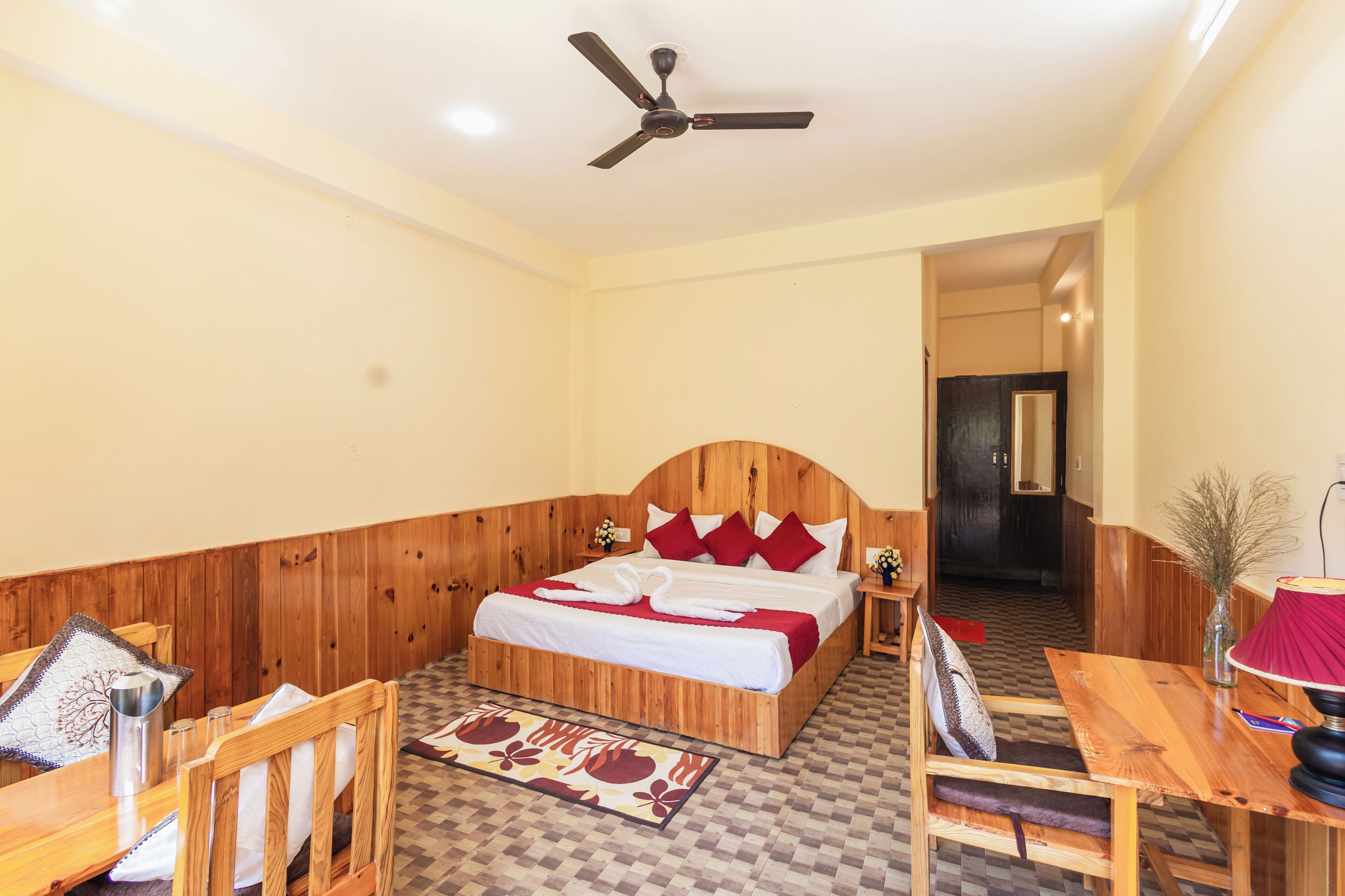 Ein Hotelzimmer mit einem hölzernen Bett, einem Esstisch, Stühlen, einem Deckenventilator und einem karierten Boden.