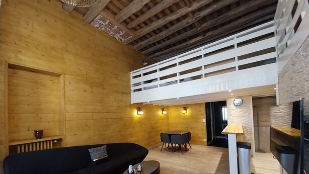 Un interior moderno con un sofá seccional negro, una mesita redonda, una zona de comedor con una mesa pequeña y sillas, y un espacio tipo loft en altura.