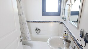 Ein Badezimmer mit Badewanne, Toilette und Waschbecken. Über der Badewanne befindet sich ein Fenster und über dem Waschbecken ein Spiegel mit Beleuchtung.