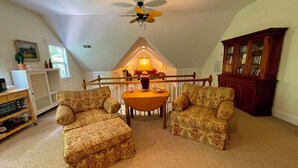 Ein gemütliches Dachzimmer mit zwei gemusterten Sesseln, einem Holzespeisetisch, einem Deckenventilator und einer Holzkabine mit Glastüren.