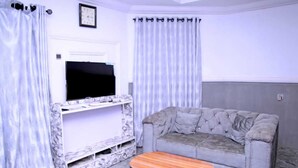 Una sala de estar con un sofá gris, una mesa de centro de madera, un televisor de pantalla plana sobre una consola de mármol y cortinas claras.