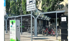 Un espace de stationnement pour vélos abrité, équipé d’une borne de recharge et d’un panneau indiquant un service de location de vélos.