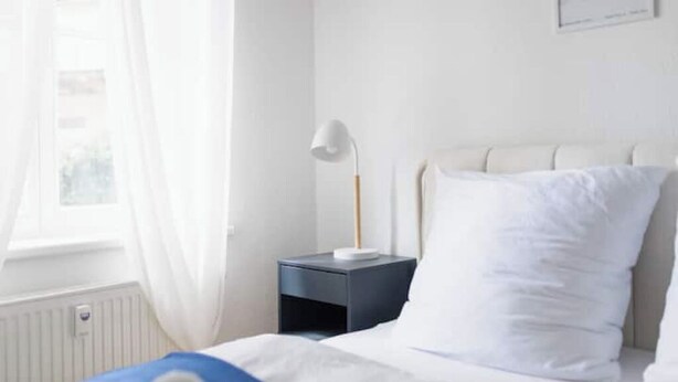 Un lit bien fait, avec une literie blanche et bleue, une lampe blanche posée sur une table de chevet sombre, et une fenêtre ornée de rideaux légers.