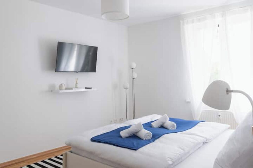 Ett hotellrum med en säng, en tv som är monterad på väggen, en sänglampa och ett fönster med tunna, genomskinliga gardiner.