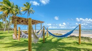 A hammock tied between wooden posts on a grassy area with a wooden pergola and palm trees in the background. - ORLA VILLAS - SÃO MIGUEL DOS MILAGRES (Passo de Camaragibe)
