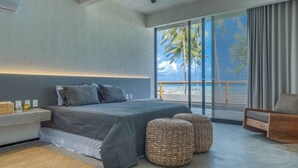 A modern bedroom with a large bed, a nightstand, wicker ottomans, and a view of the beach through large windows. - ORLA VILLAS - SÃO MIGUEL DOS MILAGRES (Passo de Camaragibe)