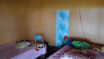 Quarto pequeno, com pouca luz, com duas camas de solteiro, uma cadeira e um prateleira fixada na parede.