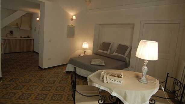 A hotel room with a bed, bedside lamps, a small table with a lamp, a decorative basket, and a view of a kitchen area. - Lavender, apartment in the heart of the historic center  (Martina Franca)