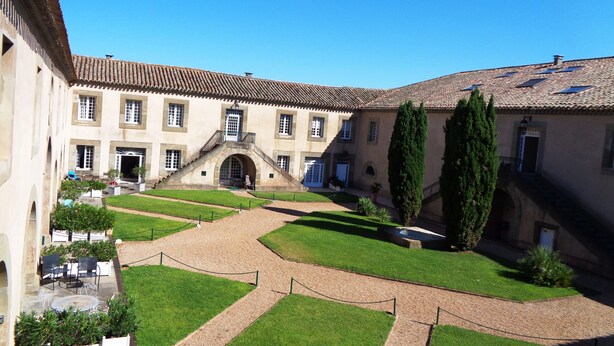 Une cour intérieure avec un escalier central, entourée d’un bâtiment doté de nombreuses fenêtres et d’un toit recouvert de tuiles.