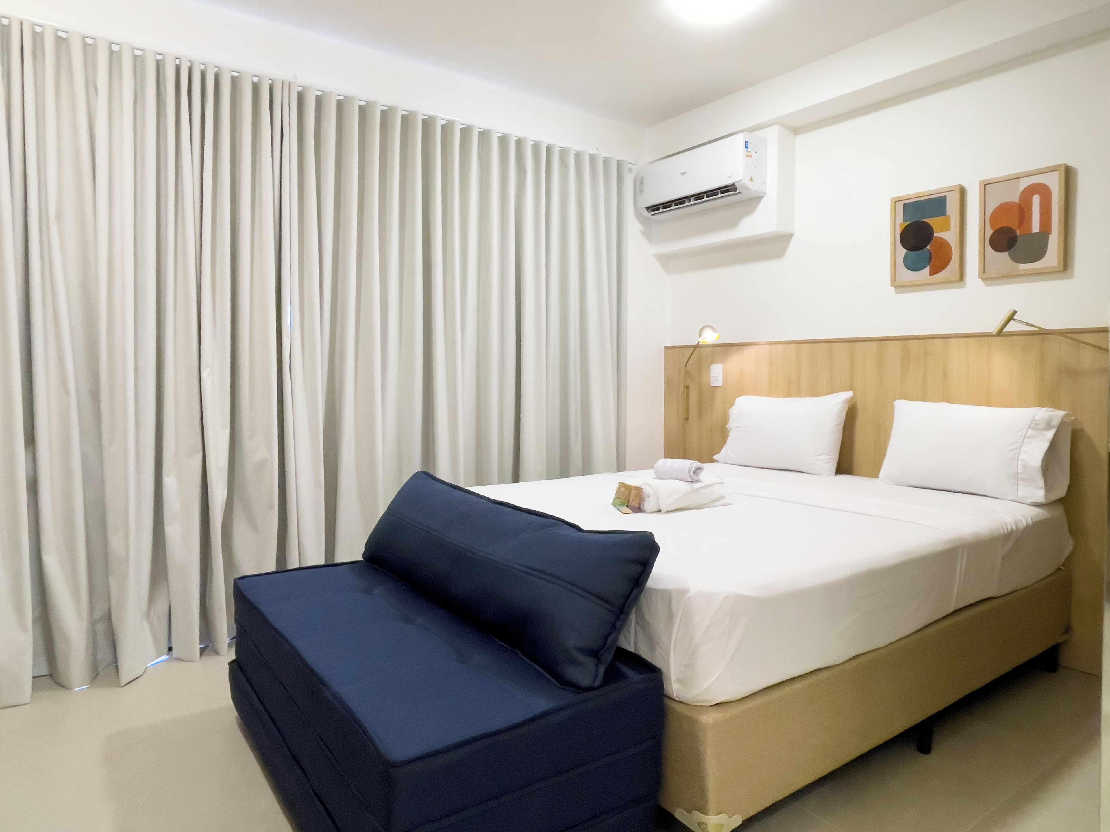 Ein Hotelzimmer mit einem Bett, einem Nachttisch, einer Lampe, einem Vorhangfenster und einer Klimaanlage.