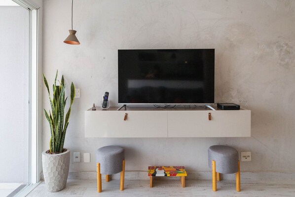Una sala de estar moderna con un televisor montado en un mueble blanco, una planta en maceta y dos taburetes grises.