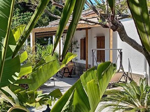 A white building with a wooden door and windows, surrounded by lush greenery.