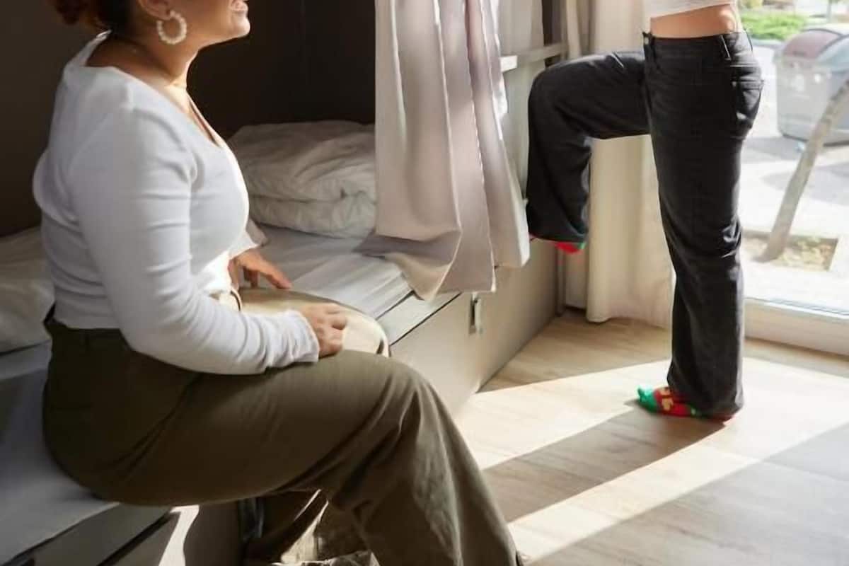 A bunk bed room with a person sitting on the lower bunk and another person standing by the window.