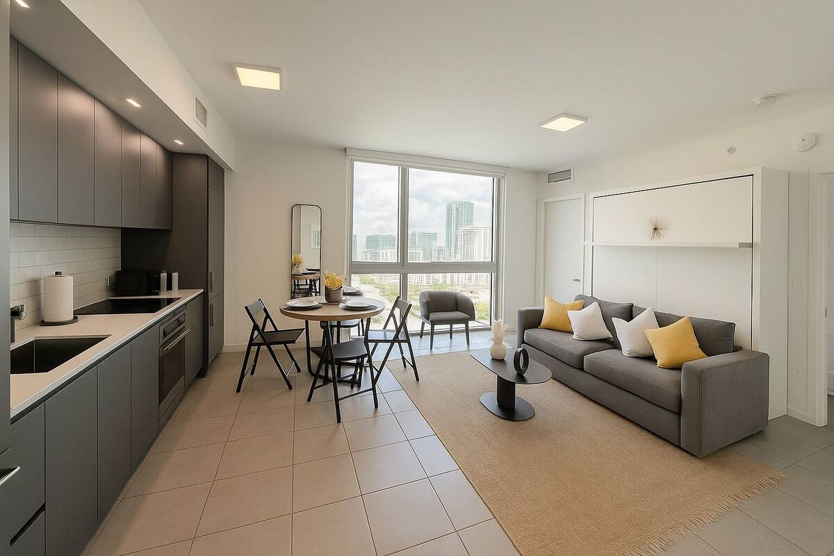 A modern apartment with a kitchen, dining area, and living room. It features a large window, a gray sofa, and a dining table with chairs.
