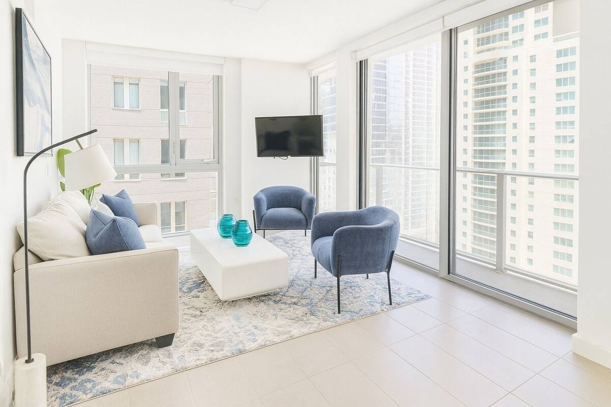 A modern living room with a sofa, two armchairs, a coffee table, and a flat-screen TV. Large windows offer a view of a cityscape.